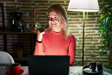 Canvas Print - Blonde woman using laptop at night at home smiling with happy face looking and pointing to the side with thumb up.