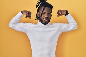 Poster - African man with dreadlocks wearing turtleneck sweater over yellow background showing arms muscles smiling proud. fitness concept.