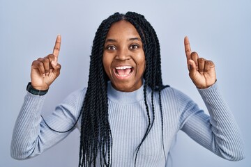 Sticker - African american woman standing over blue background smiling amazed and surprised and pointing up with fingers and raised arms.