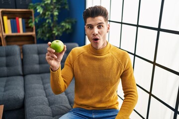 Sticker - Young hispanic man holding green apple scared and amazed with open mouth for surprise, disbelief face