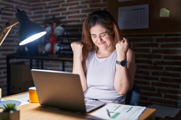 Poster - Brunette woman working at the office at night very happy and excited doing winner gesture with arms raised, smiling and screaming for success. celebration concept.