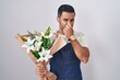 Hispanic man with beard working as florist smelling something stinky and disgusting, intolerable smell, holding breath with fingers on nose. bad smell