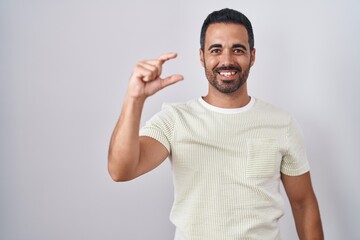 Poster - Hispanic man with beard standing over isolated background smiling and confident gesturing with hand doing small size sign with fingers looking and the camera. measure concept.