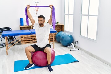 Canvas Print - Young hispanic man patient having rehab session using fit ball and elastic band at clinic