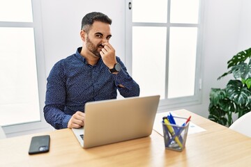 Sticker - Young hispanic man with beard working at the office with laptop smelling something stinky and disgusting, intolerable smell, holding breath with fingers on nose. bad smell