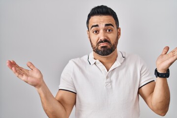 Poster - Young hispanic man with beard wearing casual clothes over white background clueless and confused expression with arms and hands raised. doubt concept.