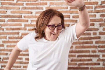 Wall Mural - Senior woman with glasses standing over bricks wall dancing happy and cheerful, smiling moving casual and confident listening to music