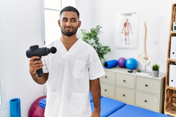 Poster - Young indian physiotherapist holding therapy massage gun at wellness center smiling looking to the side and staring away thinking.