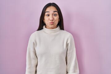 Sticker - Young south asian woman standing over pink background puffing cheeks with funny face. mouth inflated with air, crazy expression.