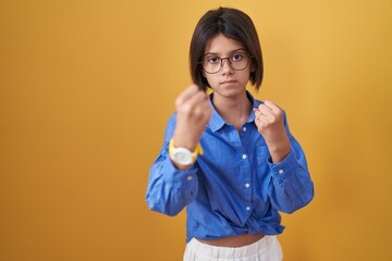 Young girl standing over yellow background ready to fight with fist defense gesture, angry and upset face, afraid of problem