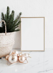 Christmas interior composition with  blank vertical picture frame mockup. Xmas Tree branches in the basket and golden christmas decorations. White wall and marble table  background. Empty copy space. 