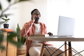 Wide shot of happy, smiling African American black young man talking on the phone while working at home office.