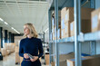 © Westend61 - Blond businesswoman holding virtual reality simulator standing by rack at warehouse