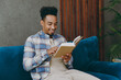 © ViDi Studio - Young student fun man wear casual shirt sits on blue sofa couch read novel stories book stay at home hotel flat rest relax spend free spare time in living room indoors grey wall People lounge concept