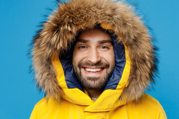 Young smiling happy fun cool satisfied man 20s wearing yellow puffer down jacket with fur hood looking camera isolated on plain blue color background studio portrait. People winter lifestyle concept.