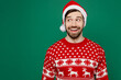 © ViDi Studio - Merry smiling happy fun young man 20s wear red warm knitted sweater Santa hat posing look aside on workspace isolated on plain dark green background studio. New Year 2023 holiday celebration concept.