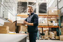 Cheerful Senior Man Smiling While Fulfilling Orders In A Warehouse. Mature Warehouse Worker Packing Goods Into Cardboard Boxes