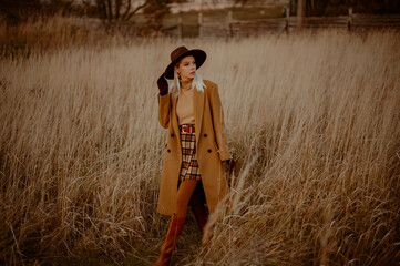 Elegant fashionable blonde woman wearing trendy autumn outfit with classic brown coat, hat, beige turtleneck, tights, mini plaid skirt, leather high boots, walking outdoor. Copy, empty space for text
