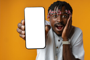 Excited young african man holding smartphone with white blank screen in hand on yellow studio background