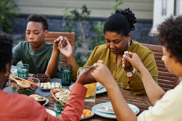 Wall Mural - Portrait of black young woman saying grace at table outdoors during family gathering and holding hands