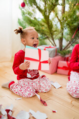 Children unpacking Christmas gifts. African American girl in Xmas pajamas sitting on the floor near the christmas tree and opening Christmas presents on morning. Selective focus