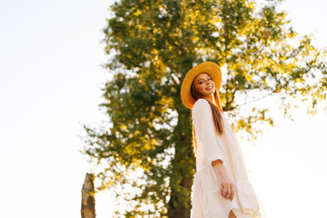 Wall Mural - low-angle view of cheerful young woman wearing straw hat and white dress standing posing on beautifu