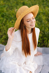 Wall Mural - vertical portrait of smiling redhead young woman sitting on a blanket on green grass relaxing at loc
