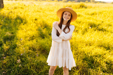 Portrait of elegant happy redhead woman in straw hat and white dress standing posing with closed eyes on beautiful meadow of green grass, on background of bright sunlight at summer day.