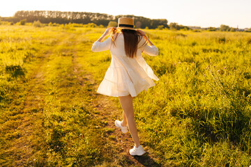 Wall Mural - back view of unrecognizable young woman in white dress holding straw hat walking at green countrysid