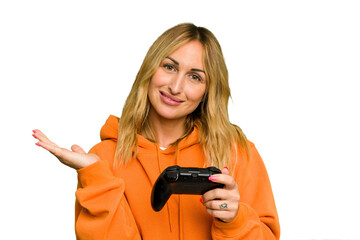 Wall Mural - Young caucasian gamer woman holding a game controller isolated on green chroma background showing a copy space on a palm and holding another hand on waist.