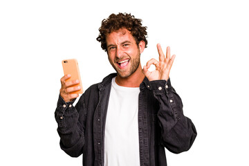 Young caucasian man using mobile phone isolated cheerful and confident showing ok gesture.