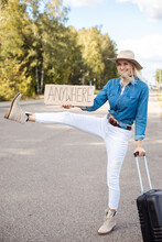 Joyful Woman With Suitcase Try To Stop Passing Car With Outstretched Raised Leg And Cardboard Poster On Empty Highway. Lady In Hat Escape From City To Go Anywhere. Travelling, Hitchhiking, Vacations