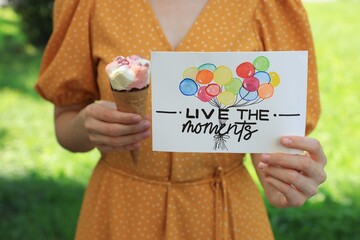 Woman holding card with phrase Live The Moments and ice cream outdoors, closeup