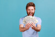 © khosrork - Portrait of extremely happy bearded man holding lot of money and screaming to camera, showing dollar bills in hand, enjoying financial success. Indoor studio shot isolated on blue background.
