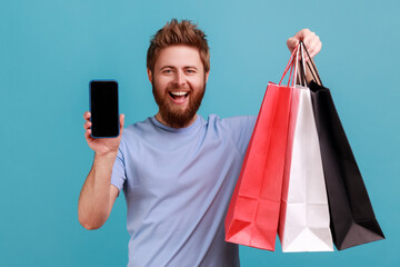 Wall Mural - portrait of optimistic bearded man standing with satisfied look showing shopping bags and smart phon