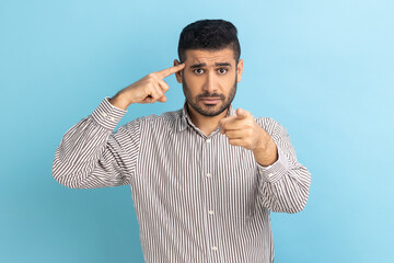 Businessman twirling finger to temple pointing finger at camera, saying you are dumb and stupid, dissatisfied with dumbness, wearing striped shirt. Indoor studio shot isolated on blue background.