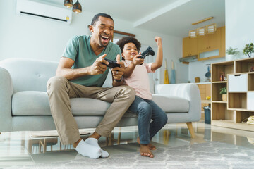 Wall Mural - Cheerful Black African American Father and his Little son in Afro hair sitting on sofa using joysticks or game controllers playing console video games at home