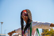 an African American woman with long sisterlocks wearing white and pink clothes, sunglasses and an orange head scarf carrying and green purse doing yoga poses on the rocks of a river 