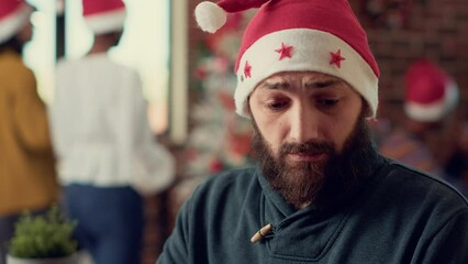 Canvas Print - Startup employee working on laptop during winter holiday, planning report in office with christmas tree decorations. Young man wearing santa hat and celebrating festivity. Close up. Handheld shot.