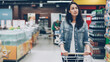© silverkblack - Pretty young lady is walking through aisle in spacious supermarket with shopping cart looking at shelves with products, employees in uniform are working in background.