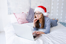 Caucasian Young Woman While Working On Laptop At Home For Christmas In Cozy Clothes And A Santa Hat Sits On A Bed In A Decorated Bedroom With A Christmas Tree. New Year And Christmas Concept.