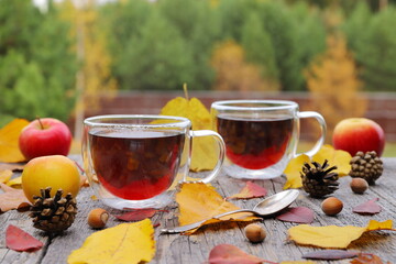 Canvas Print - A glass mugs of hot tea are among foliage, fruit, cones and nuts on a wooden table on the autumn forest background.