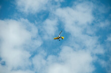 A Yellow Motorized Hang Glider With A Propeller Soars In The Blue Sky Against The Background Of Clouds