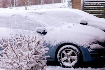 A portrait of two cars covered in white snow parked in a driveway. The driver will have to clear all windows from snow or wait for the snow to melt in order to start driving safely.