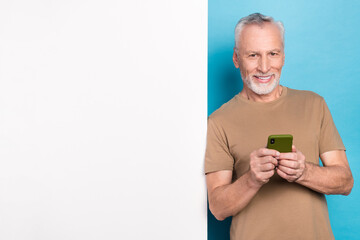 Sticker - Portrait of cheerful retired man with gray hair beard beige t-shirt hold smartphone lean on touch screen isolated on blue color background