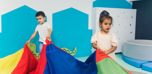 Wall Mural - two preschoolers holding a big fabric and trying to play with it, medium shot kindergarten kids' concept. High quality photo