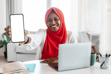 Poster - Business app. African american muslim woman showing smartphone with blank screen, sitting at desk with laptop, mockup