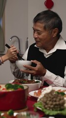 Wall Mural - Vertical Screen: happy asian grandfather talking with hand gestures as his polite granddaughter putting food into his bowl at dinner table on chinese lunar new year's eve at home