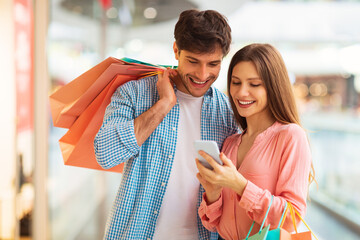 Poster - Couple Shopping Online Using Smartphone Posing Holding Bags In Mall
