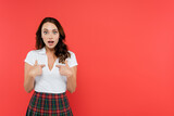 Fototapeta Na ścianę - Amazed woman in plaid skirt pointing with fingers isolated on red